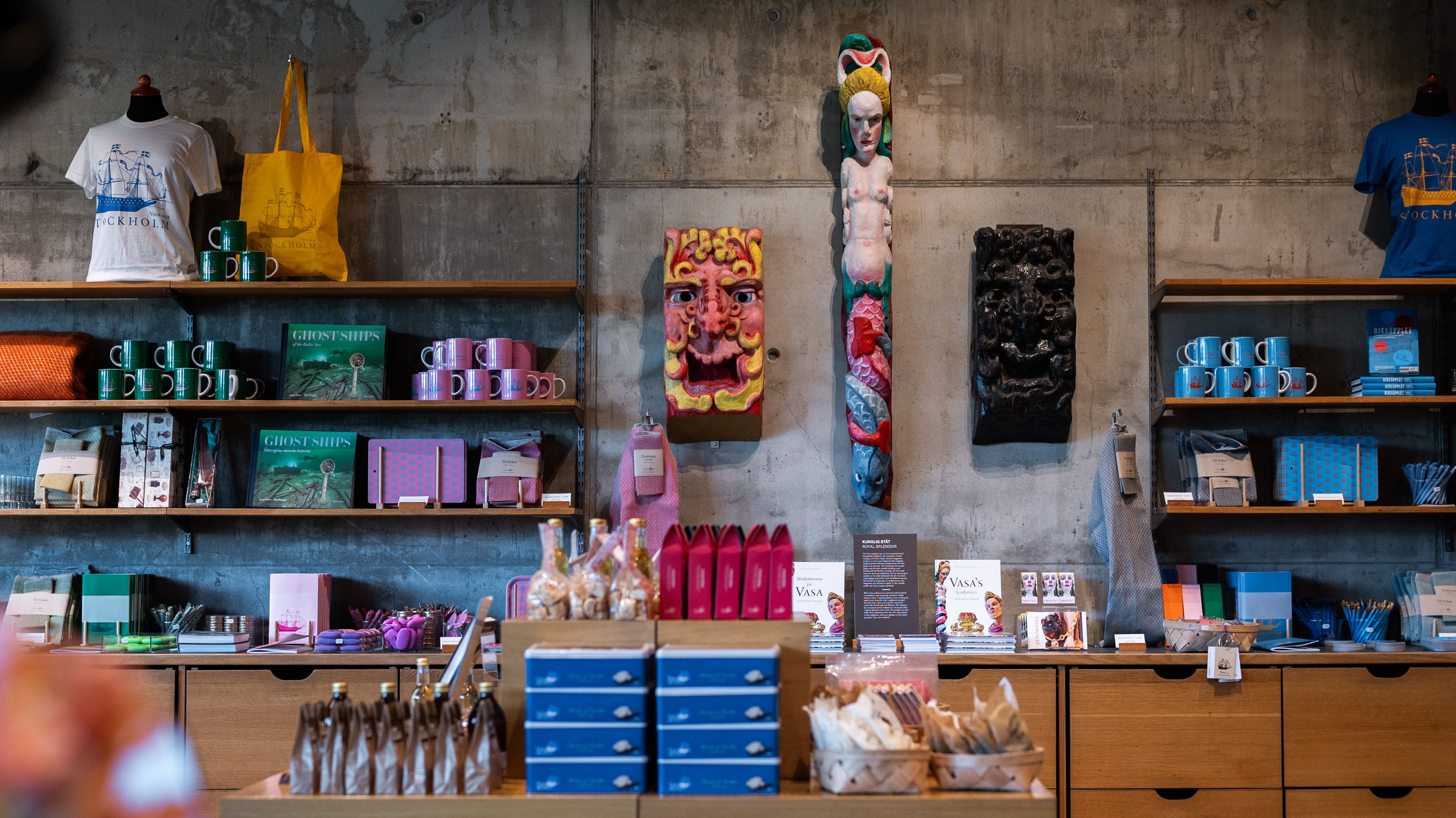Foto av vägg i Vasamuseets butik, en grå vägg med skulpturer i klara färger, hyllor med muggar, böcker och textiler. 