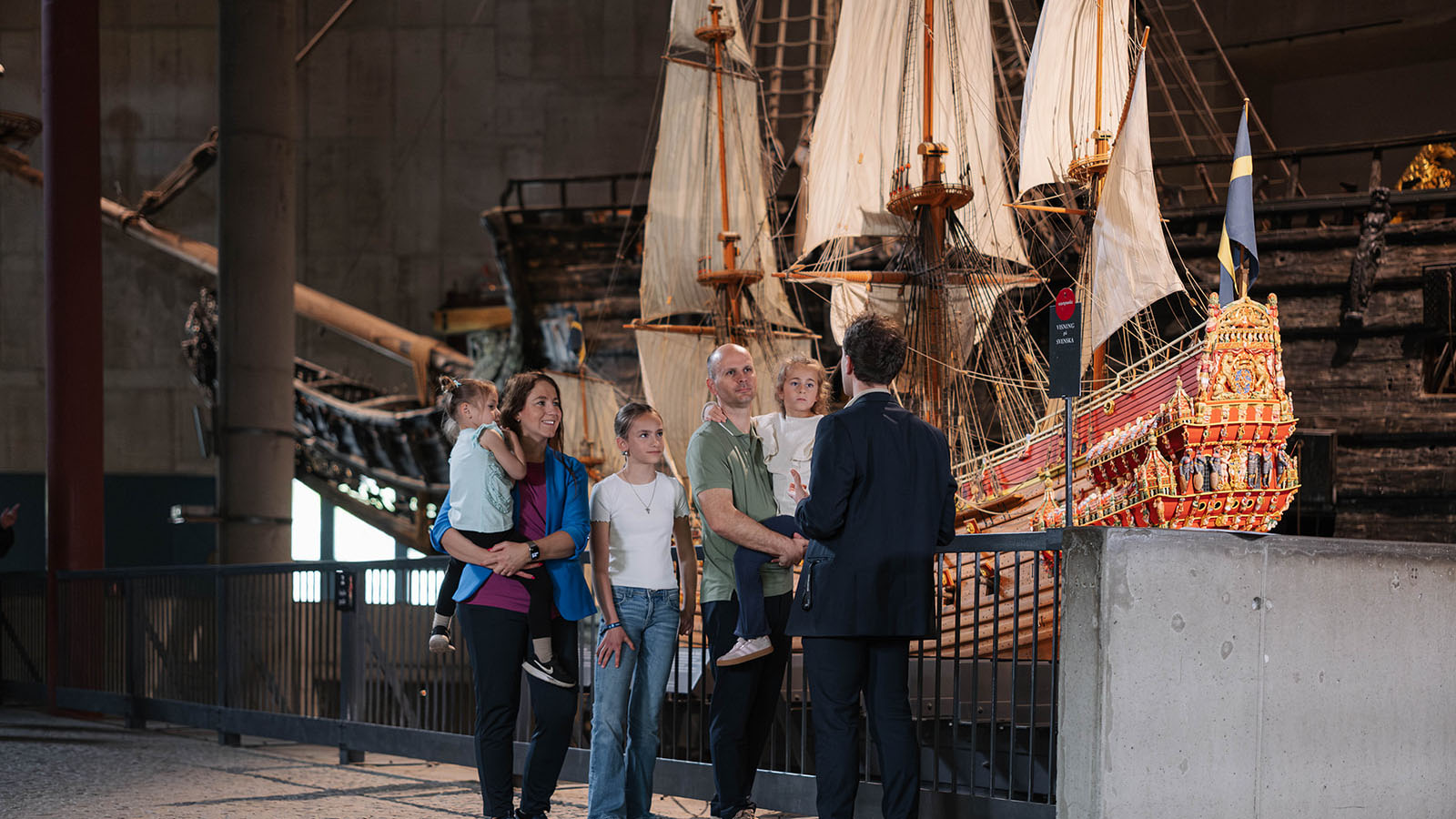 Familj med tre döttrar framför modell av skeppet Vasa, med det riktiga skeppet i bakgrunden. En guide i mörk kavaj står vänd mot familjen. 